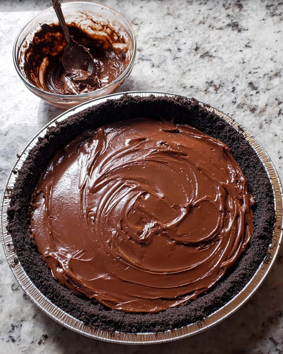 The image shows a chocolate pie with two visible layers. The bottom layer is a dark, crumbly crust that forms the edge and base of the pie. The top layer is a thick, shiny, smooth chocolate filling spread evenly with swirl marks on the surface. To the right of the pie, there is a small clear glass bowl with some leftover chocolate and a spoon inside it. The pie and bowl are placed on a white marbled textured surface. Photo taken with an iphone --ar 4:5 --v 7