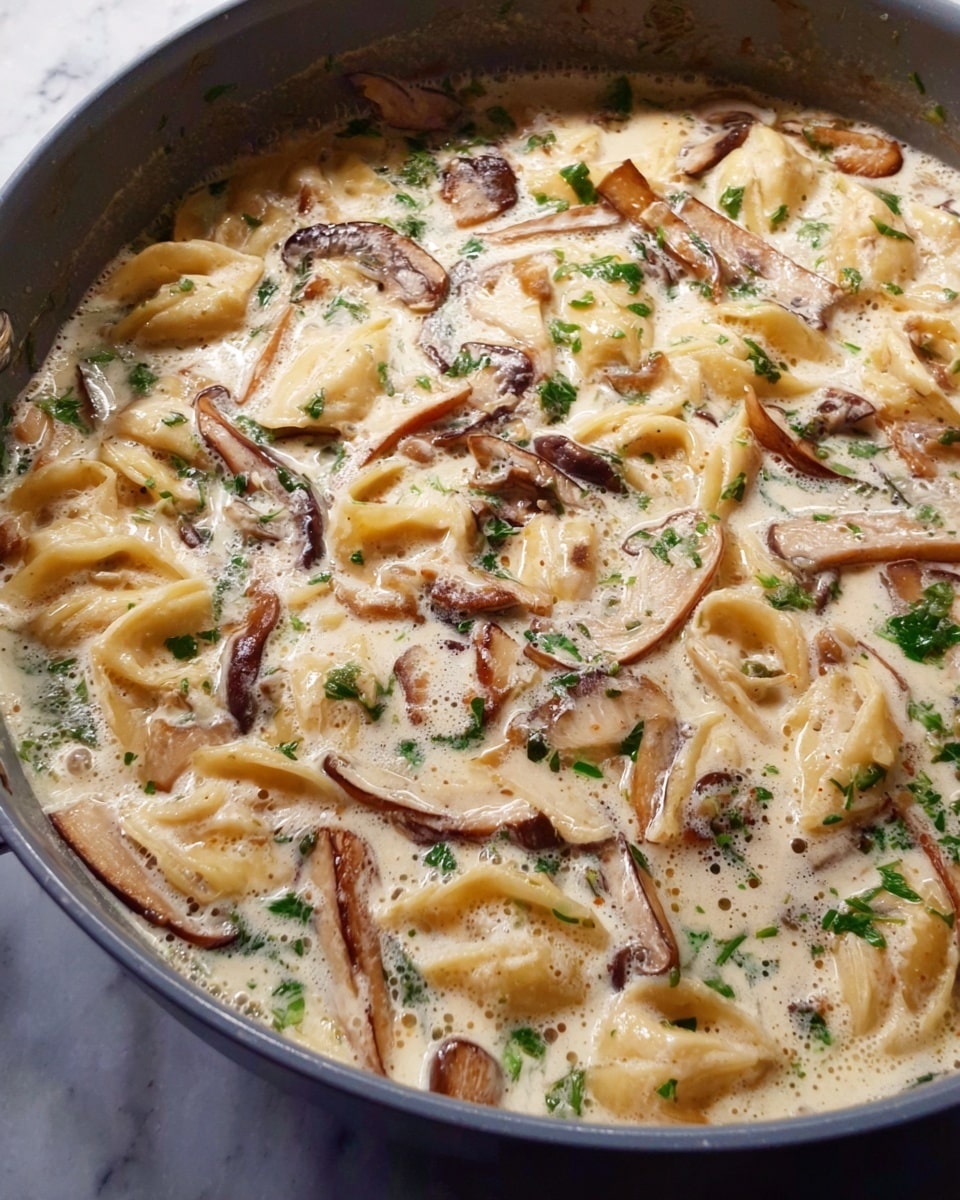 A close-up view of a creamy pasta dish in a gray pan, showing soft, slightly curved pasta pieces mixed evenly in a thick, pale cream sauce. Thin, brown mushroom slices are spread throughout the dish, adding a rich color contrast. Small green herb bits are scattered on top, giving a fresh touch to the creamy texture. The sauce looks smooth with tiny bubbles and light swirls, covering every part of the pasta and mushrooms. The pan sits on a white marbled surface. photo taken with an iphone --ar 4:5 --v 7