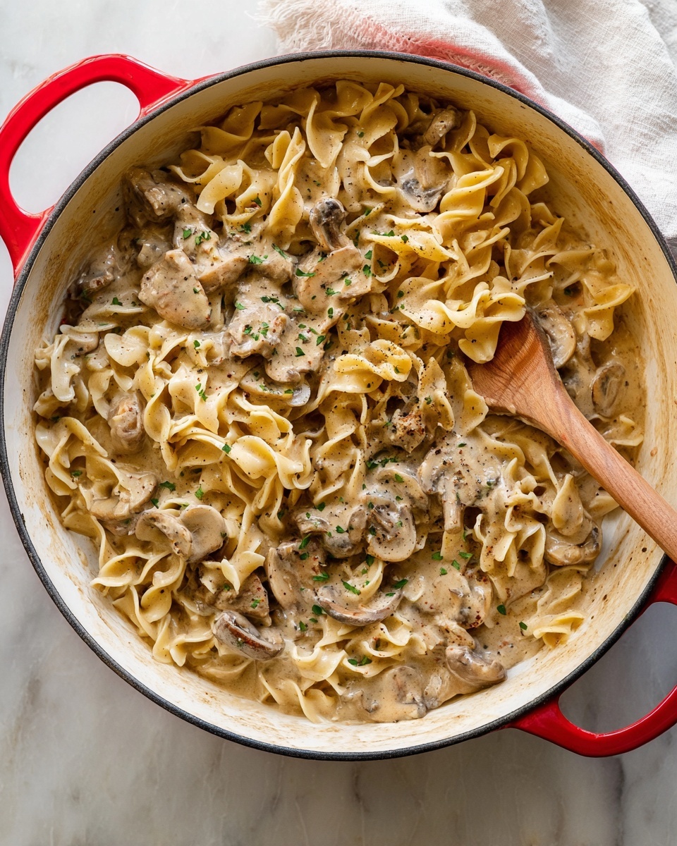 A white deep pan with red handles holds creamy pasta mixed with sliced mushrooms and pieces of cooked meat, all covered in a thick light brown sauce with specks of black pepper and green herbs scattered on top. The pasta is pale yellow with a slightly wavy texture, and the sauce coats every piece, pooling slightly at the bottom edges of the pan. A wooden spoon is partially submerged on the right side, nestled among the creamy pasta. The background is a white marbled surface with a blurred white cloth in the upper right corner. Photo taken with an iphone --ar 4:5 --v 7