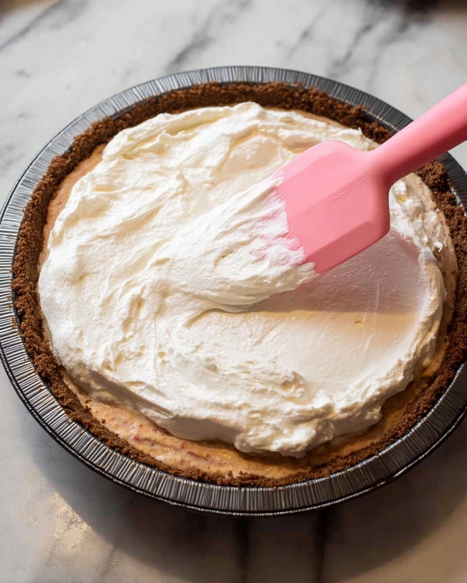 A round pie with a dark brown crust sits in a shiny metal pie tin on a white marbled surface, topped with a thick, creamy white layer of whipped topping being spread by a pink spatula. The pie filling under the topping is a rich, soft brown with a slightly uneven texture, visible around the edges where the whipped topping is not yet spread. The whipped topping is fluffy and smooth, covering about half of the pie’s surface as the pink spatula moves across it. Photo taken with an iphone --ar 4:5 --v 7