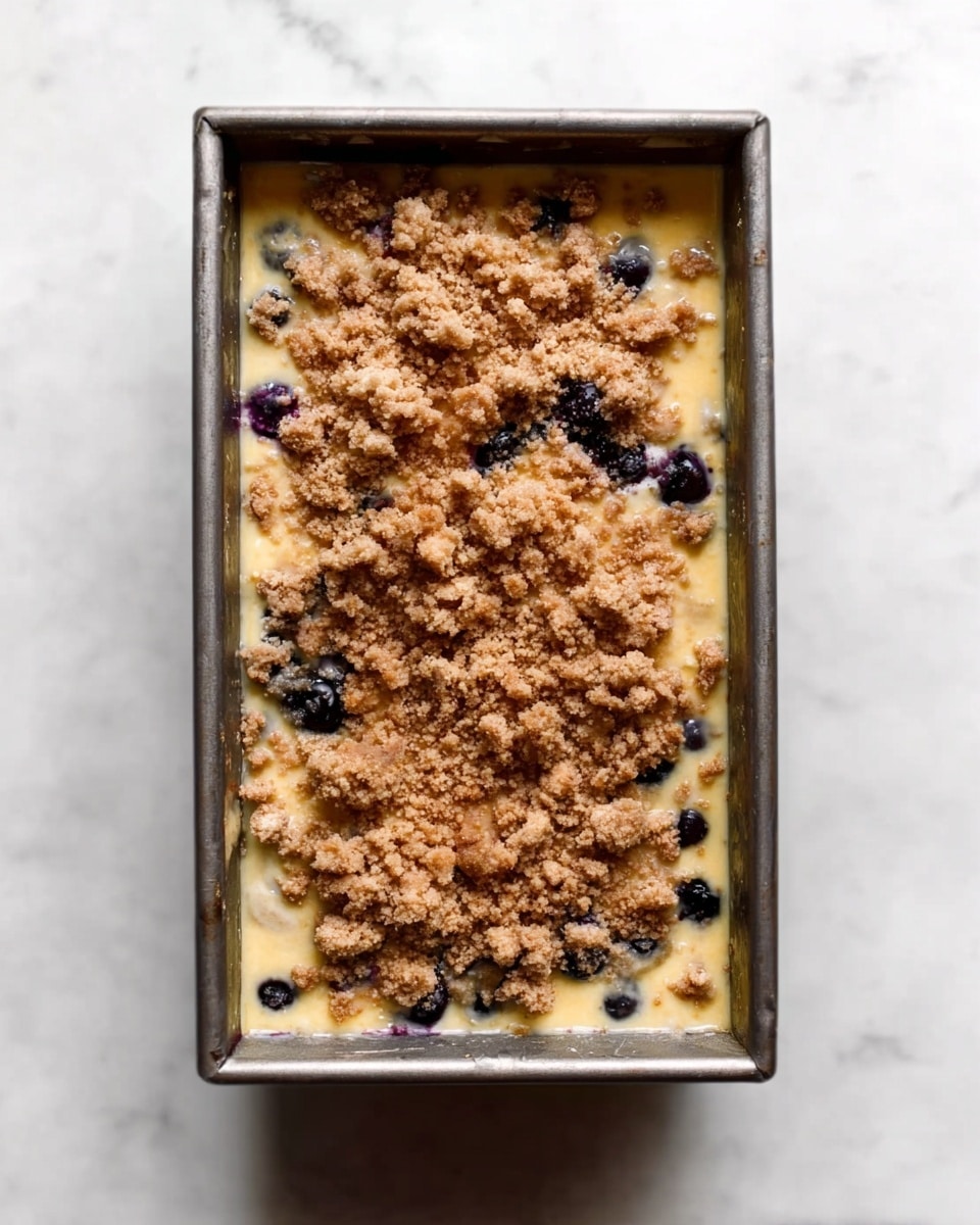 The image shows a rectangular metal baking pan with a layered dessert inside, placed on a white marbled surface. The bottom layer is a pale yellow custard-like mixture. Scattered throughout this layer are dark blueberries, some visible on the surface. On top of this is a crumbly brown topping, unevenly spread, with chunks of different sizes and a rough texture. The pan is viewed from above, showing the full rectangular shape and consistent layers. Photo taken with an iphone --ar 4:5 --v 7