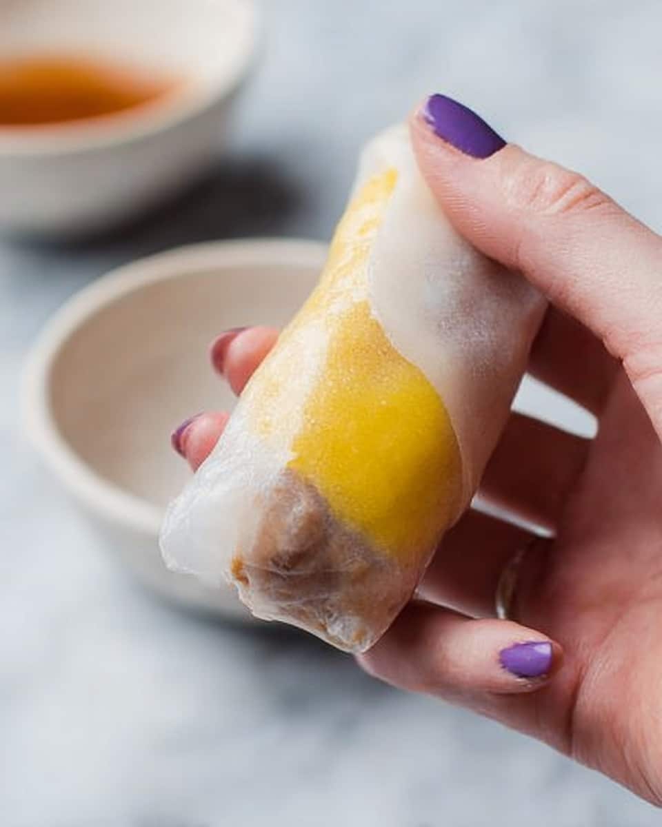 A close-up of a woman's hand holding a translucent spring roll showing two clear layers inside, one brown and one bright yellow. The spring roll looks soft and slightly wet, with the woman's fingers gently pinching it near the bottom. In the blurred background, there is a simple white bowl on a white marbled surface. The woman's nails are painted purple. photo taken with an iphone --ar 4:5 --v 7