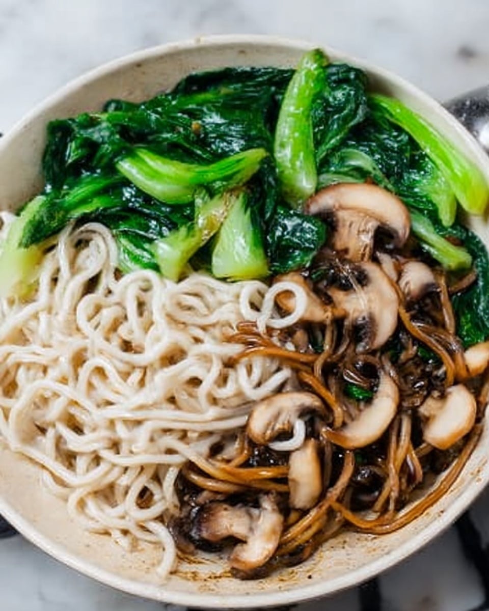A close-up of a dish in a white bowl featuring white and brown noodles mixed together in the center, surrounded by cooked green leafy vegetables and sliced mushrooms. The green leaves are bright and soft, while the mushrooms are light brown and slightly glossy. The noodles are smooth and piled loosely, with the brown noodles coated in sauce and the white noodles plain. The bowl sits on a white marbled surface. Photo taken with an iphone --ar 4:5 --v 7