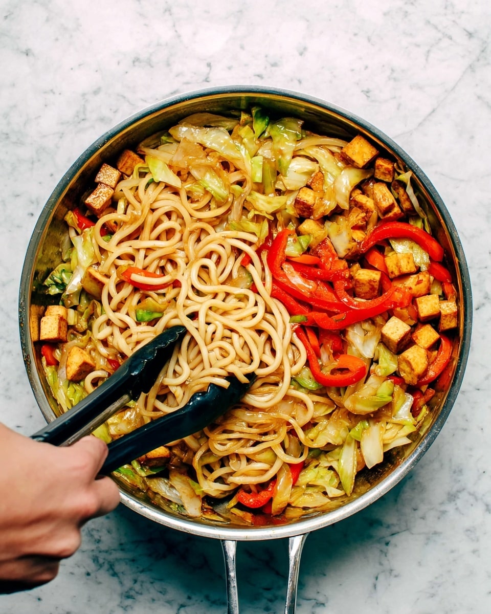 A metal pan filled with a colorful stir-fry dish sits on a white marbled surface. Inside the pan, there are three main layers: at the bottom, light green and pale yellow cabbage pieces mixed with slices of green vegetables; above that, curly pale beige noodles tangled throughout; and on top, small golden-brown tofu cubes and bright red strips of bell pepper scattered evenly. A woman's hand holds black tongs grabbing some noodles and vegetables in the pan. The mix looks fresh and well cooked with a slightly shiny texture. Photo taken with an iphone --ar 4:5 --v 7