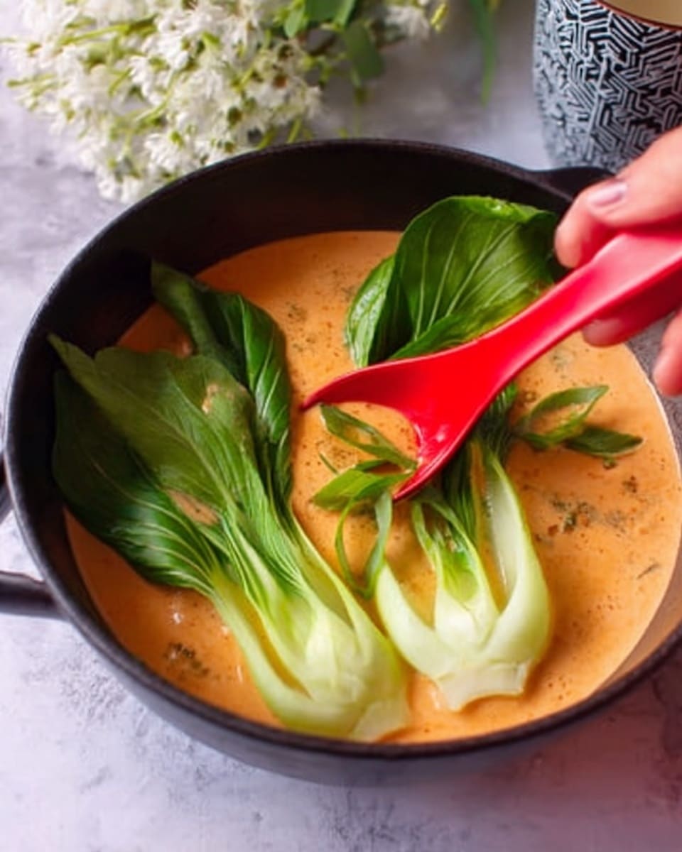 The image shows a black bowl filled with a creamy orange soup that has a smooth texture. Inside the soup, there are three whole green bok choy leaves placed on the right side, standing upright and partially submerged. A vibrant red spoon is held by a woman's hand on the left side, stirring the soup gently. The bowl sits on a white marbled surface with a small bunch of white flowers and a patterned cup visible softly blurred in the background. photo taken with an iphone --ar 4:5 --v 7