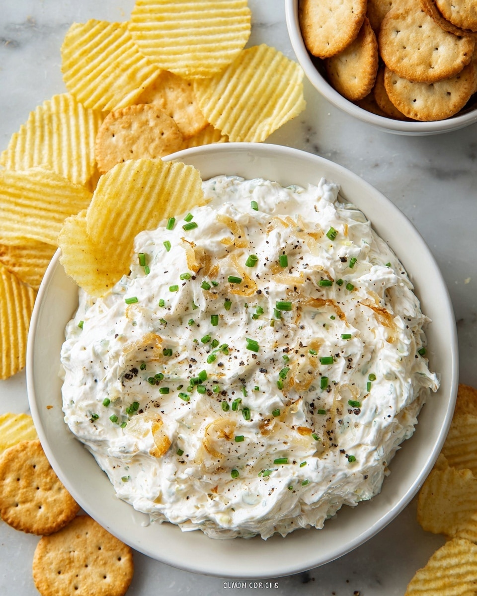 A white plate holds a creamy dip with a thick texture, mostly white with small green chive pieces scattered evenly on top, along with golden-brown cooked onions and light specks of black pepper. The dip looks thick and slightly chunky. Around the plate on a white marbled surface, there are light yellow ridged potato chips and a white bowl filled with round light brown crackers. The chips and crackers are casually arranged, some chips stacked in the bowl and some loose on the surface. photo taken with an iphone --ar 4:5 --v 7