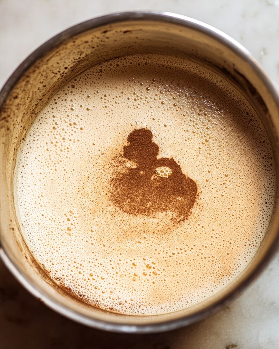A close-up view of a metal bowl filled with a light beige foamy mixture with many small bubbles visible on the surface; in the center, there is a splash of darker brown powder creating an irregular shape with fine particles spreading slightly outward; the inside of the bowl shows some residue and condensation near the rim, with the whole scene placed on a white marbled surface photo taken with an iphone --ar 4:5 --v 7