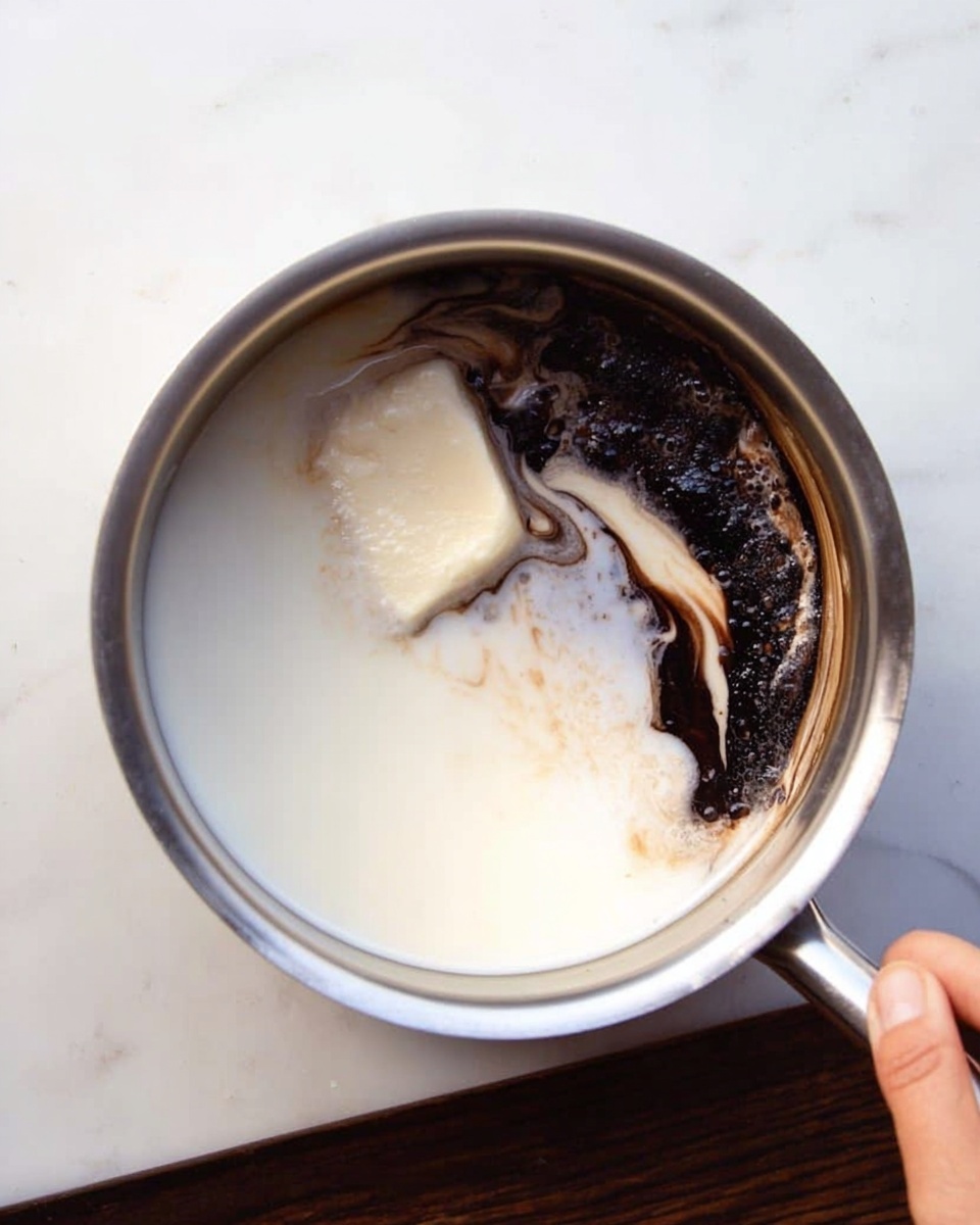 A shiny silver pot with a small amount of white liquid filling half the pot. There is a light cream solid piece floating in the liquid toward the center, and a dark brown mixture settling mostly on one side, partially mixed with the white liquid, creating a swirling effect. The pot is held by a woman's hand on the right side, and it rests on a white marbled surface. photo taken with an iphone --ar 4:5 --v 7