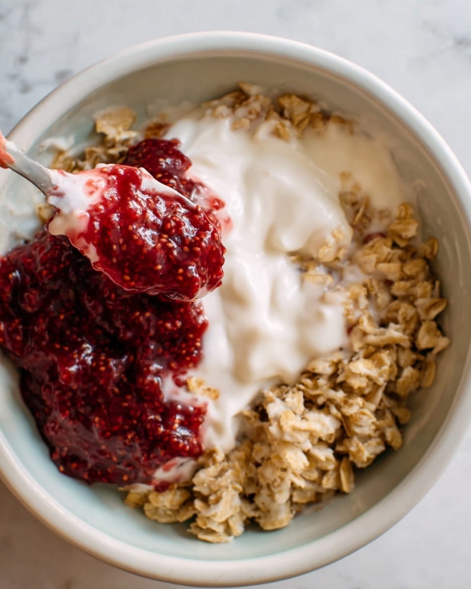 The image shows a close-up view of a bowl filled with three main layers. At the top, there is a creamy white yogurt layer with a smooth texture, partially covering the other ingredients. Next to the yogurt lies a dark red chia seed jam layer, thick and slightly chunky with visible chia seeds spread unevenly. The base layer consists of light brown oats, which appear soft and slightly wet from mixing with yogurt. A woman's hand holds a spoon in the top left corner of the bowl, scooping some mixture. The bowl is white and set on a white marbled surface. Photo taken with an iphone --ar 4:5 --v 7