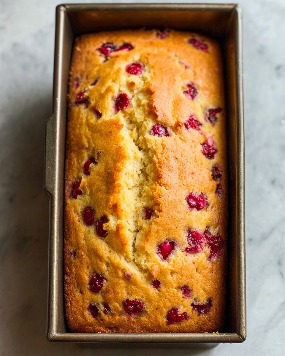 A golden brown loaf cake with small red berries evenly spread throughout the top and visible inside, sitting inside a rectangular metal baking pan. The cake has a slightly cracked texture running down the middle, showing a soft and moist inside beneath the golden crust. The pan is placed on a white marbled surface. photo taken with an iphone --ar 4:5 --v 7