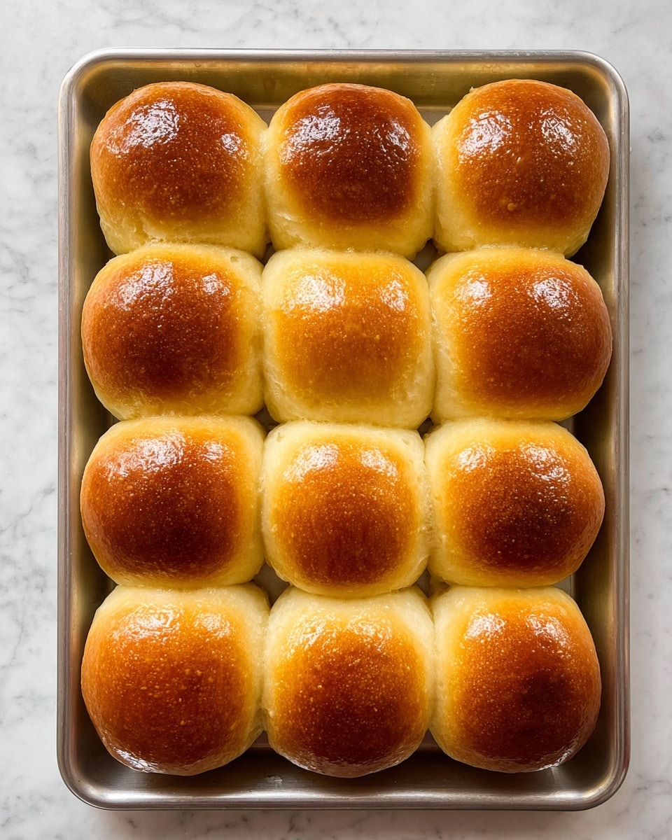 The image shows a baking tray with twelve golden brown bread rolls arranged in a 3x4 grid. Each round roll has a shiny, smooth top with a slightly darker, rich brown color, contrasting with the lighter, soft-looking sides. The rolls are touching each other, forming a connected pattern on the tray. The tray is placed on a white marbled surface. photo taken with an iphone --ar 4:5 --v 7