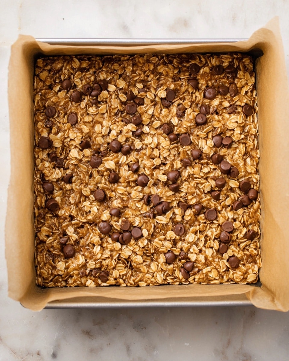 The image shows a square metal baking pan lined with light brown parchment paper filled with a thick, evenly spread oat and chocolate chip mixture. The top layer is golden brown with visible whole oats and scattered round chocolate chips embedded into the surface. The texture looks rough and chunky with small, uneven oat pieces throughout. The pan sits on a white marbled surface. Photo taken with an iphone --ar 4:5 --v 7