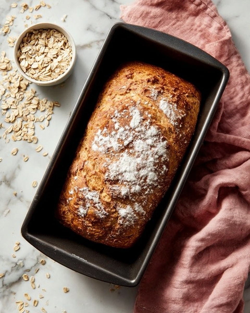 A golden-brown loaf of bread with a slightly rough, cracked surface dusted with white flour sits in a dark metal loaf pan. The bread looks soft inside with crispy edges. To the left of the loaf pan, a small white bowl holds uncooked rolled oats, and some oats are scattered lightly on the white marbled surface. A pink cloth is placed underneath the loaf pan on the right side, adding softness to the scene. photo taken with an iphone --ar 4:5 --v 7