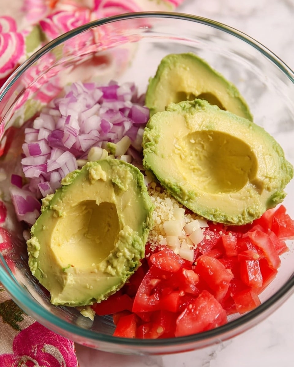 The image shows a clear glass bowl filled with three large halves of bright green avocado placed in the center. Around the avocado halves, there are three piles of finely chopped ingredients: purple-red onion pieces in the top part of the bowl, bright red tomato cubes on the right side, and a small mound of light beige minced garlic on the lower left side. The background has a white marbled texture with a pink and white floral cloth partially visible on the left side. photo taken with an iphone --ar 4:5 --v 7