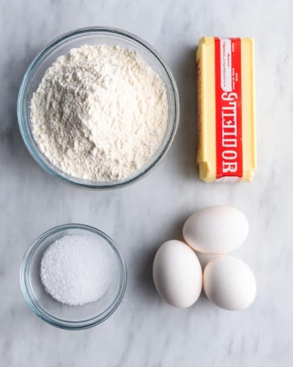 The image shows four cooking ingredients placed on a white marbled surface. In the top left corner, there is a round clear glass bowl filled with white flour. At the top right, there is a stick of yellow butter with red and white packaging. Below the flour and butter, there are three white eggs arranged together on the right side. To the left of the eggs, there is another smaller round clear glass bowl containing granulated white sugar. The overall setting is simple and clean, with the ingredients evenly spaced out. Photo taken with an iphone --ar 4:5 --v 7