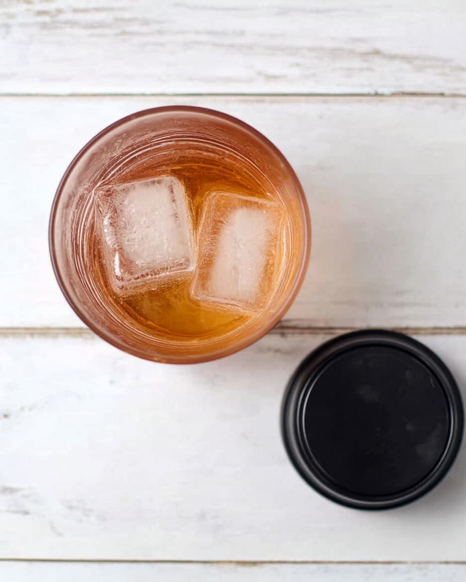 The image shows a clear glass filled with a light amber drink and two ice cubes floating on top. The glass is viewed from above. Next to the glass is a matte black bottle cap resting on a white marbled surface. The background consists of white wood planks. Photo taken with an iphone --ar 4:5 --v 7