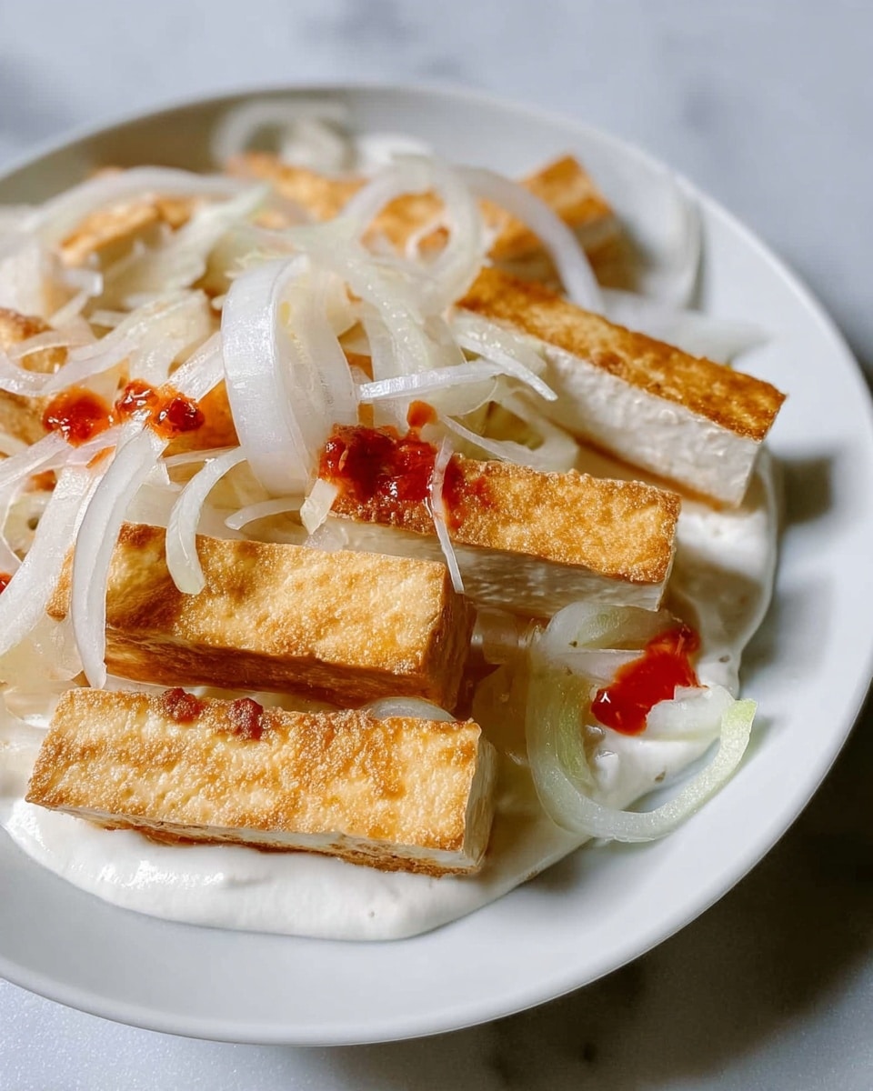The image shows a white plate with several layers of food. The bottom layer is a white creamy sauce spread over the plate. On top of this sauce, there are rectangular pieces of tofu with a golden brown crispy texture. Thinly sliced white onions are spread on and around the tofu pieces. There is also a small amount of red sauce drizzled over some of the tofu, adding a splash of color. The plate is set on a white marbled surface. Photo taken with an iphone --ar 4:5 --v 7