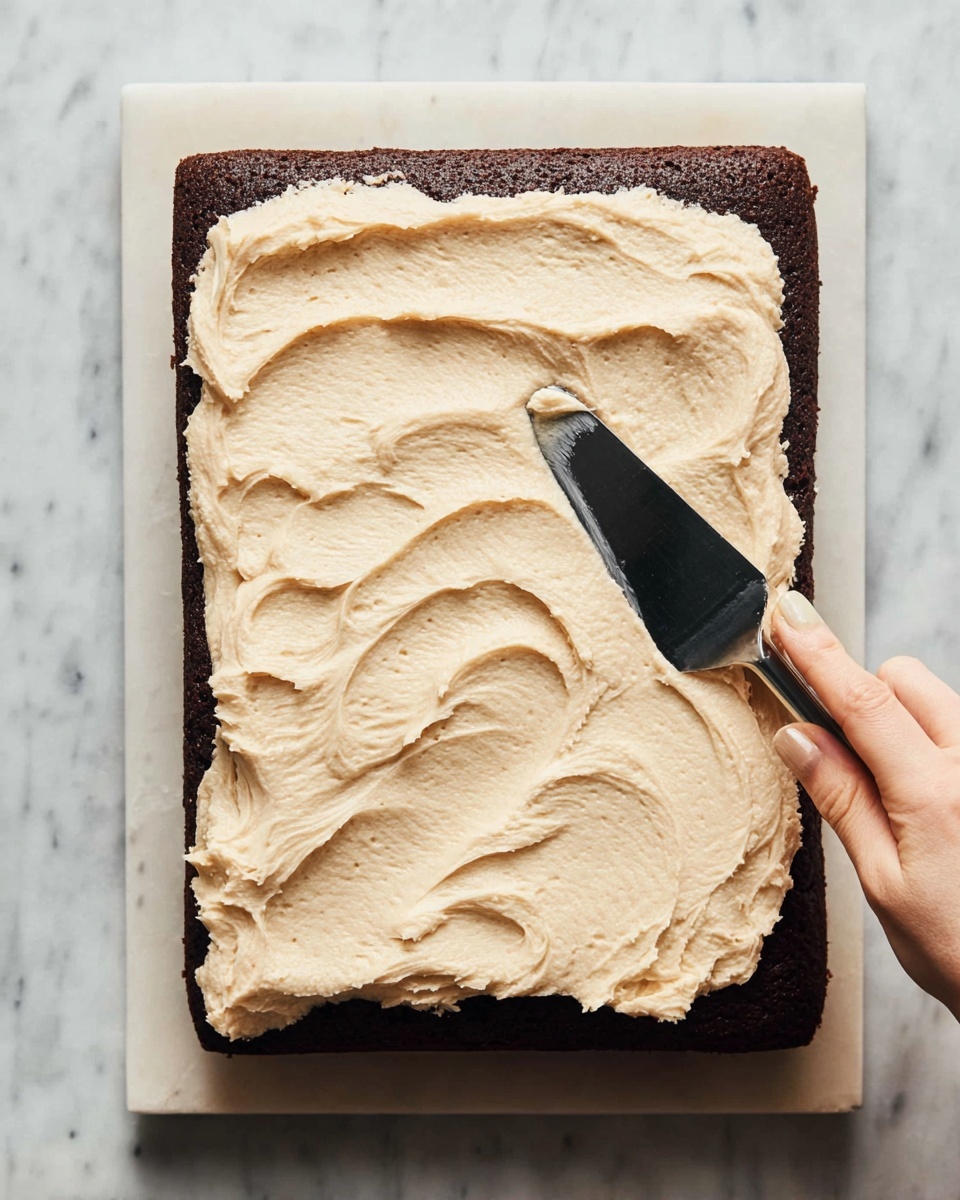 A rectangular dark brown cake sits on a white marbled surface with a thick layer of light beige frosting spread unevenly on top, showing creamy texture and soft peaks. A woman's hand holds a black-handled spatula, smoothing the frosting on the cake's center, leaving visible swirls and ridges as it spreads outward. The close-up view focuses on the contrast between the dark cake and pale frosting, highlighting the rich texture of both. photo taken with an iphone --ar 4:5 --v 7