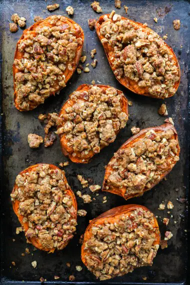 The image shows many pieces of roasted orange sweet potato with crispy edges on a gray baking tray. The sweet potato pieces are cut into irregular chunks, lightly seasoned with small green herbs and glistening with a shiny coating of oil. A large metal serving spoon with a wooden handle is scooping up some sweet potato chunks from the bottom right. The background surface is a white marbled texture. Photo taken with an iphone --ar 4:5 --v 7