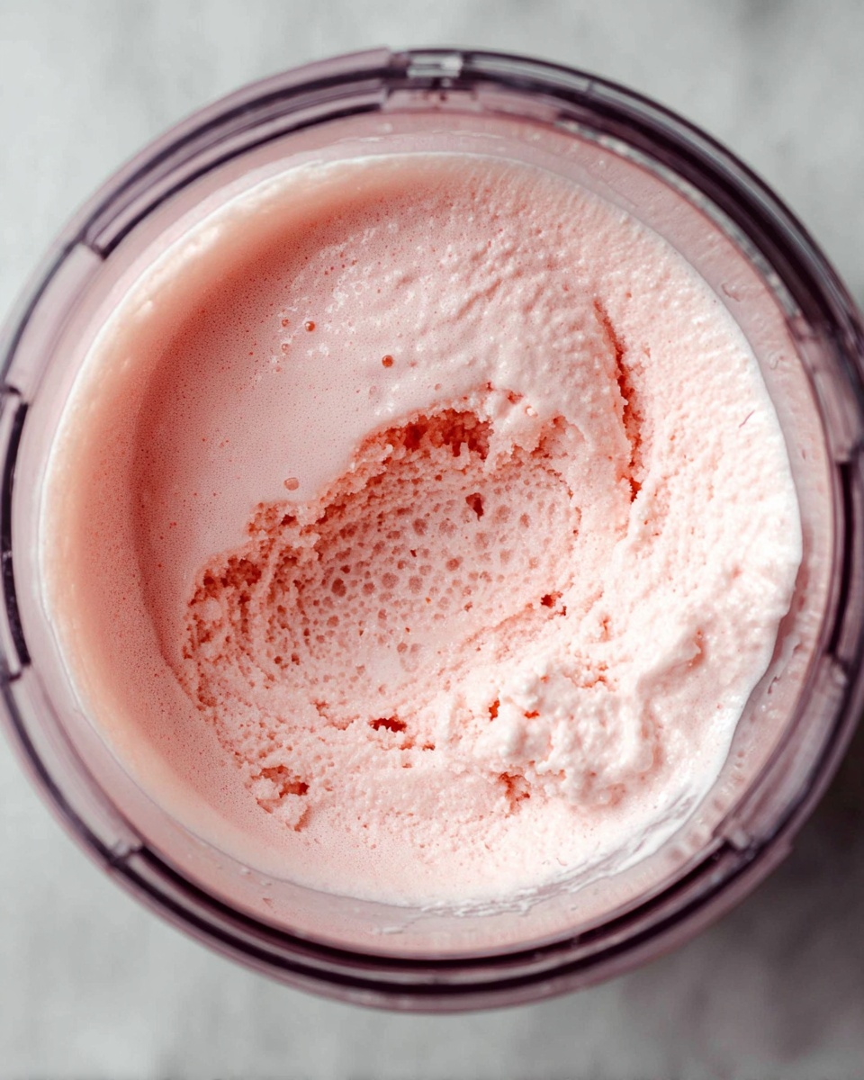 A top view of a clear container filled with a pale pink, fluffy ice cream mixture that has a soft, smooth texture on the edges and a slightly melted, bubbly liquid layer in the center, surrounded by a thick, whipped layer. The container sits on a white marbled surface, with the ice cream mixture showing a foamy and airy consistency and a light pink color throughout. Photo taken with an iphone --ar 4:5 --v 7