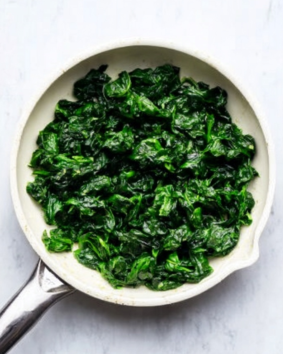 A white frying pan filled with dark green cooked chopped spinach, showing a slightly glossy texture from moisture, placed on a white marbled surface. The spinach pieces are uniformly sized and cover the whole pan bottom, with some small wrinkles and soft folds visible in the leaves. Photo taken with an iphone --ar 4:5 --v 7