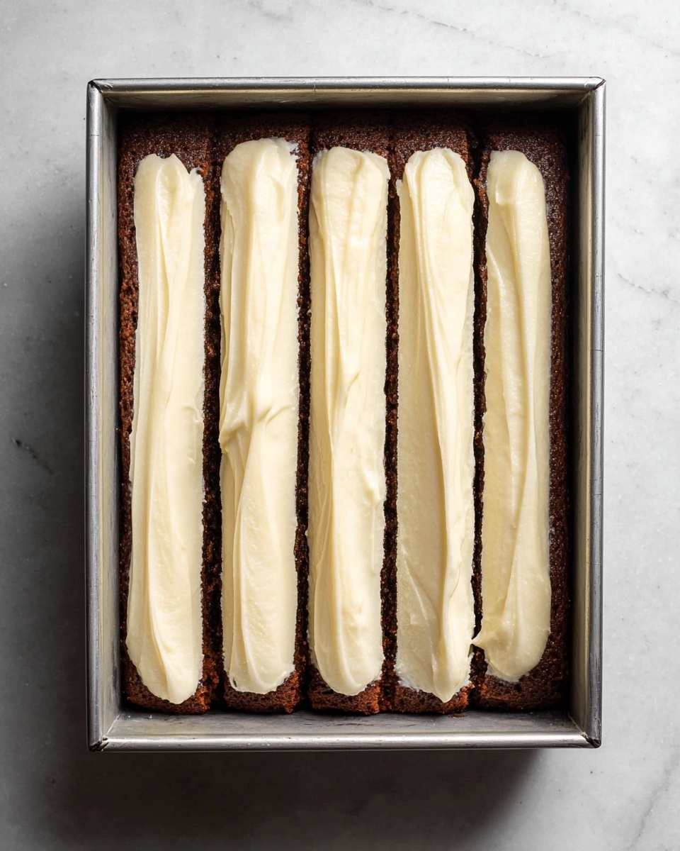 A rectangular metal pan contains a single dark brown cake layer as the base. On top of the cake layer, there are five thick, evenly spaced, long lines of creamy light beige frosting running vertically from top to bottom. The frosting lines have a smooth texture with slight swirls and are neatly placed with visible cake edges in between. The pan rests on a white marbled surface photo taken with an iphone --ar 4:5 --v 7