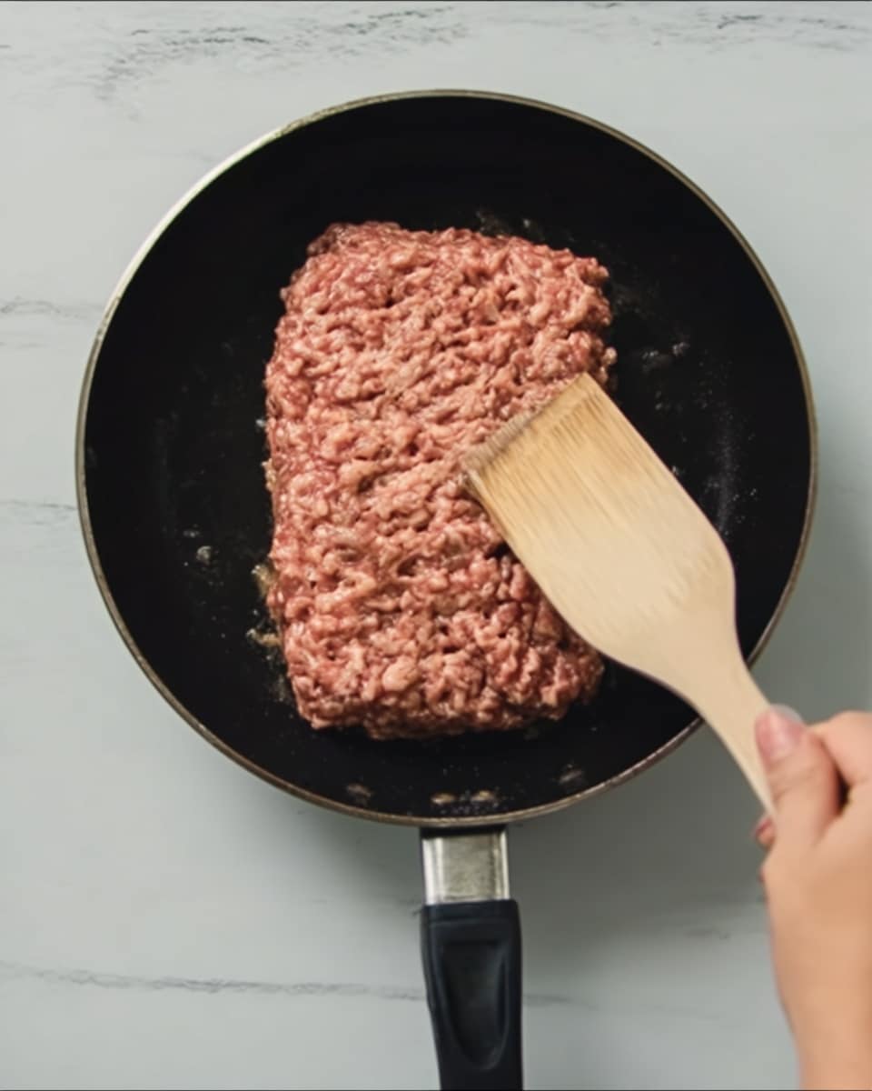 In the image, a black pan is placed on a white marbled surface. Inside the pan, there is a single layer of ground meat, which is brownish-red with a slightly uneven texture, spread flat across the pan’s bottom. A woman's hand is seen from the right side, holding a wooden spatula that is touching and pressing down on the meat. The spatula is light beige in color. The background and the pan contrast clearly, and the scene appears to be in the middle of cooking preparation. photo taken with an iphone --ar 4:5 --v 7