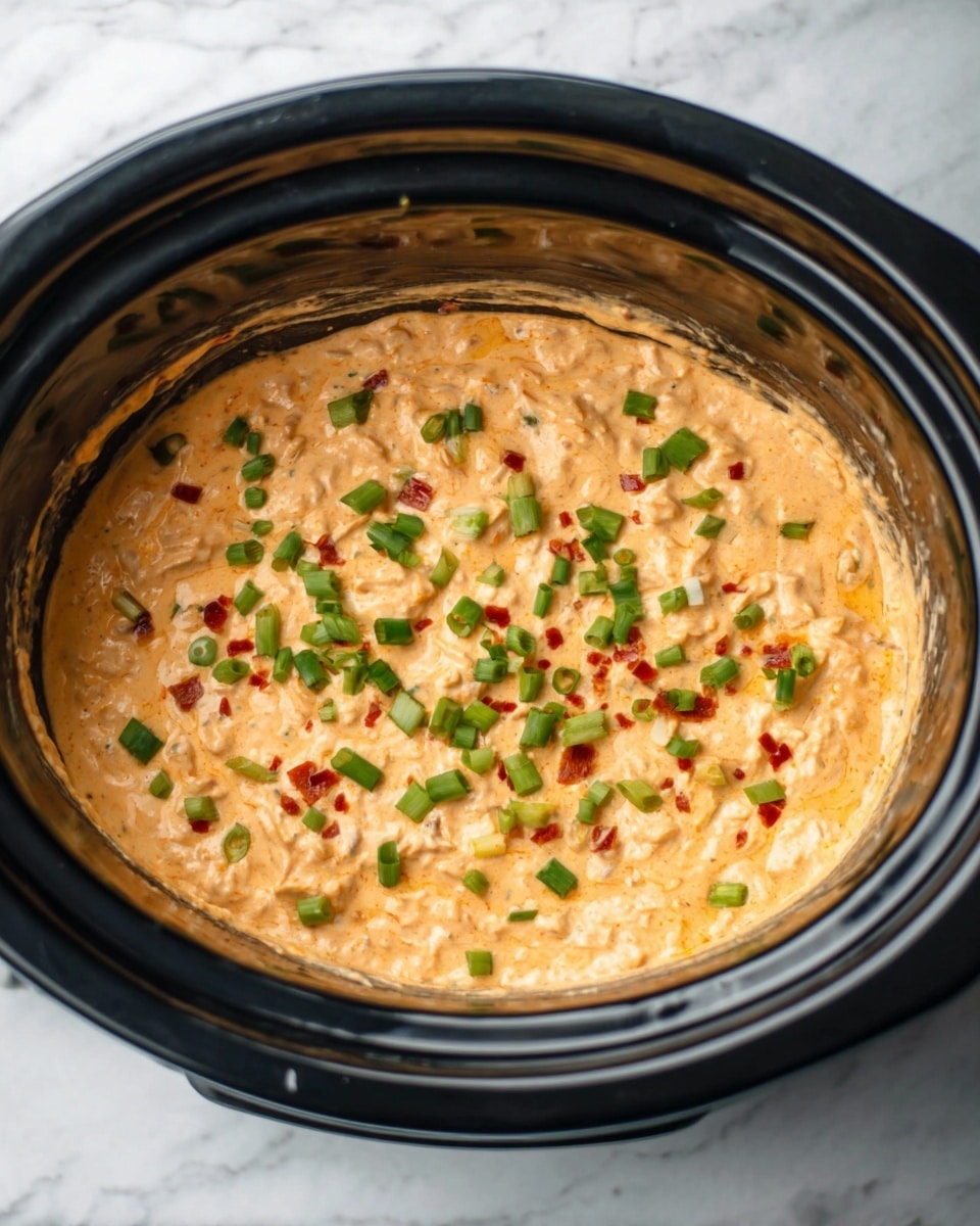 A black slow cooker is shown filled with a creamy orange dip that has small chunks inside. The dip is topped with green chopped onions and small red bits scattered evenly over the surface. The slow cooker sits on a white marbled surface. Photo taken with an iphone --ar 4:5 --v 7