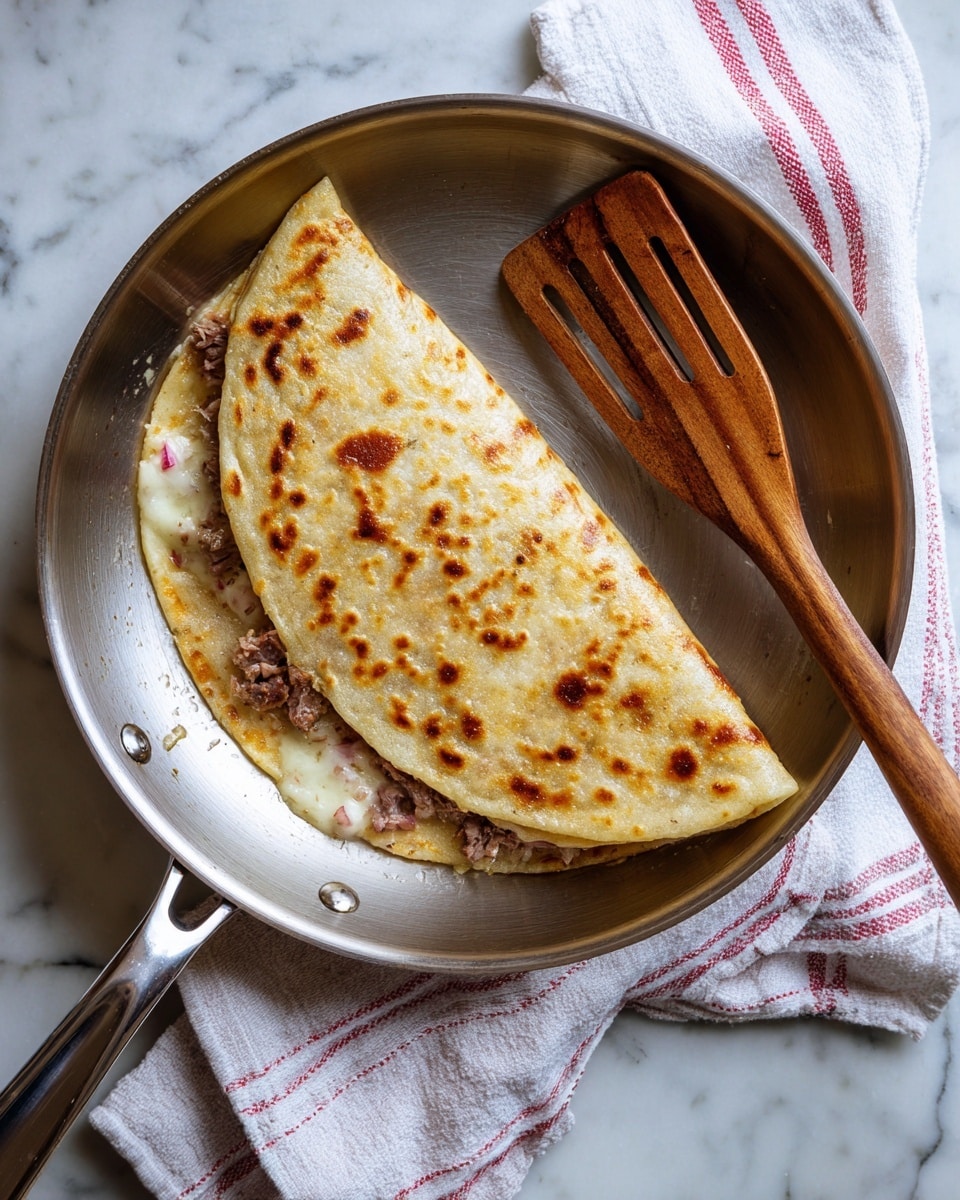 A lightly toasted folded flatbread with a golden-brown spotted surface sits inside a shiny silver pan. Inside the flatbread, a few layers of visible filling include melted white cheese, pieces of browned meat, and some small bits of red, likely tomato or pepper. A wooden spatula with slotted holes rests beside the flatbread in the pan. The pan is placed on a folded white and red striped cloth on a white marbled surface. Photo taken with an iphone --ar 4:5 --v 7