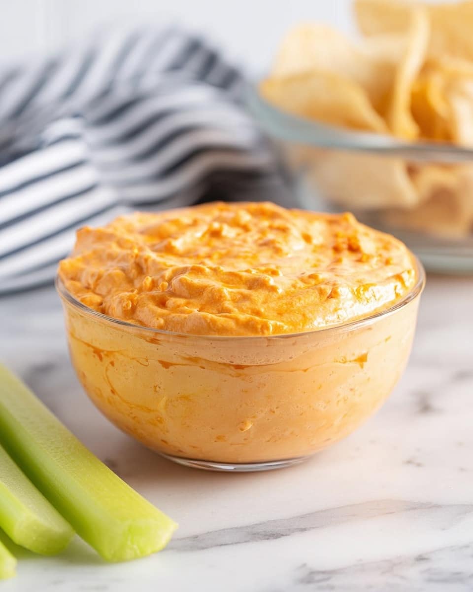 A clear glass bowl filled with a thick, creamy orange dip that has a slightly chunky texture, placed on a white marbled surface. In the background, there is a blurry glass bowl with light tan potato chips and a striped black and white cloth. In the foreground, part of three pale green celery sticks lie horizontally, partially blurred. The dip fills the glass bowl almost to the top, with a smooth but textured surface photo taken with an iphone --ar 4:5 --v 7