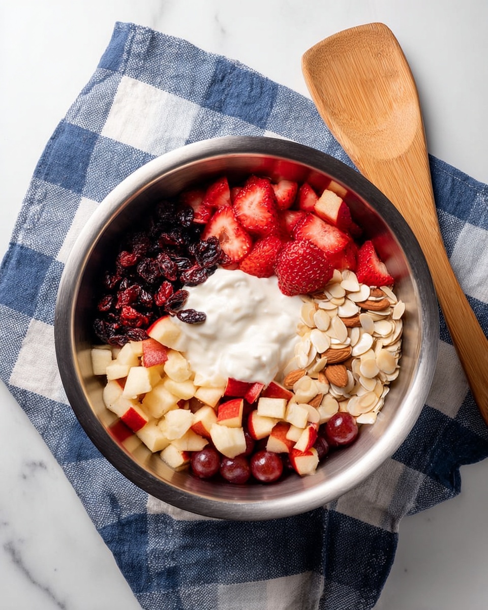 A metal bowl filled with layers of various fruits and yogurt sits on a blue and white checkered cloth on a white marbled surface. The bowl contains a center layer of white yogurt. Surrounding the yogurt, there are sliced red strawberries at the top, dark dried cranberries on the upper left, light beige sliced almonds on the upper right, red apple chunks on the lower left, and small red grapes on the lower right. A wooden spatula rests next to the bowl. Photo taken with an iphone --ar 4:5 --v 7