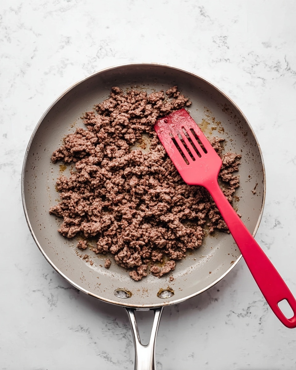 A gray pan with a shiny metal handle is placed on a white marbled surface. Inside the pan, there is a layer of browned ground meat, cooked and spread evenly across the bottom. A bright red spatula rests on the right side of the pan, partially under the meat, showing some oil or juice next to it. The image shows just one main layer of meat in the pan. photo taken with an iphone --ar 4:5 --v 7