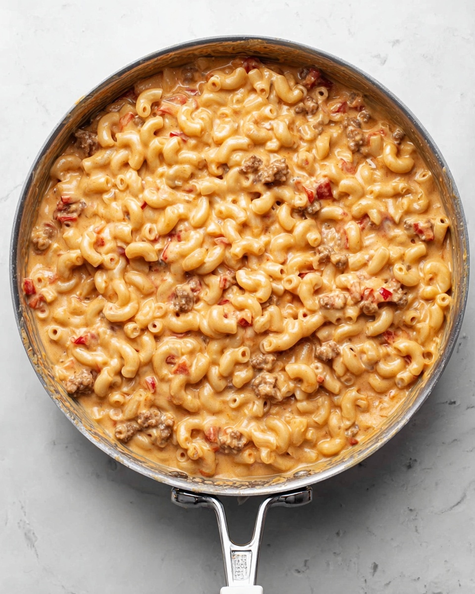 A large silver pan filled with creamy macaroni pasta mixed with small pieces of cooked meat and tiny red bits, all coated in a thick light orange sauce. The pasta is ring-shaped and covers the entire surface of the pan, with the sauce evenly spread throughout, giving a smooth and slightly glossy texture. The pan handle is silver with a white grip, placed on a white marbled surface. photo taken with an iphone --ar 4:5 --v 7