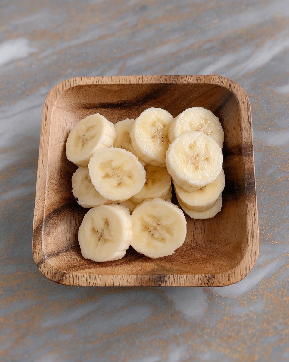 A small wooden rectangular bowl filled with about a dozen banana slices, each slice showing a creamy pale yellow color with a soft texture and small brown spots in the center, all stacked casually inside the bowl. The bowl rests on a white marbled surface. Photo taken with an iphone --ar 4:5 --v 7