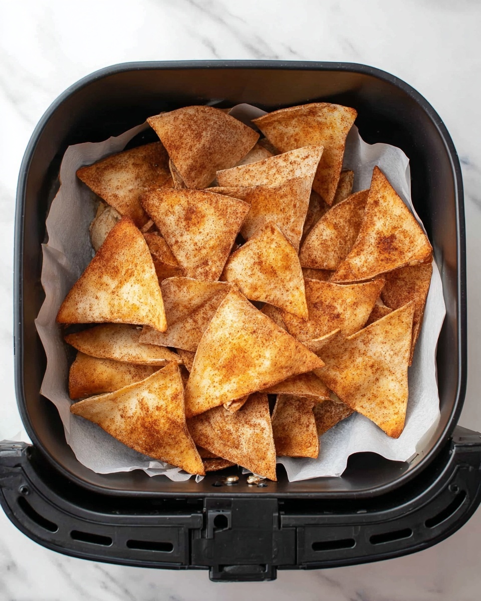 The image shows a black air fryer basket lined with white parchment paper holding many triangular chips. The chips are golden brown with a dusting of cinnamon or similar spice, giving them a slightly textured surface with spots of darker brown. The chips are layered, overlapping each other in a piled arrangement inside the basket, which has small rectangular holes around the sides. The basket is placed on a white marbled surface. photo taken with an iphone --ar 4:5 --v 7