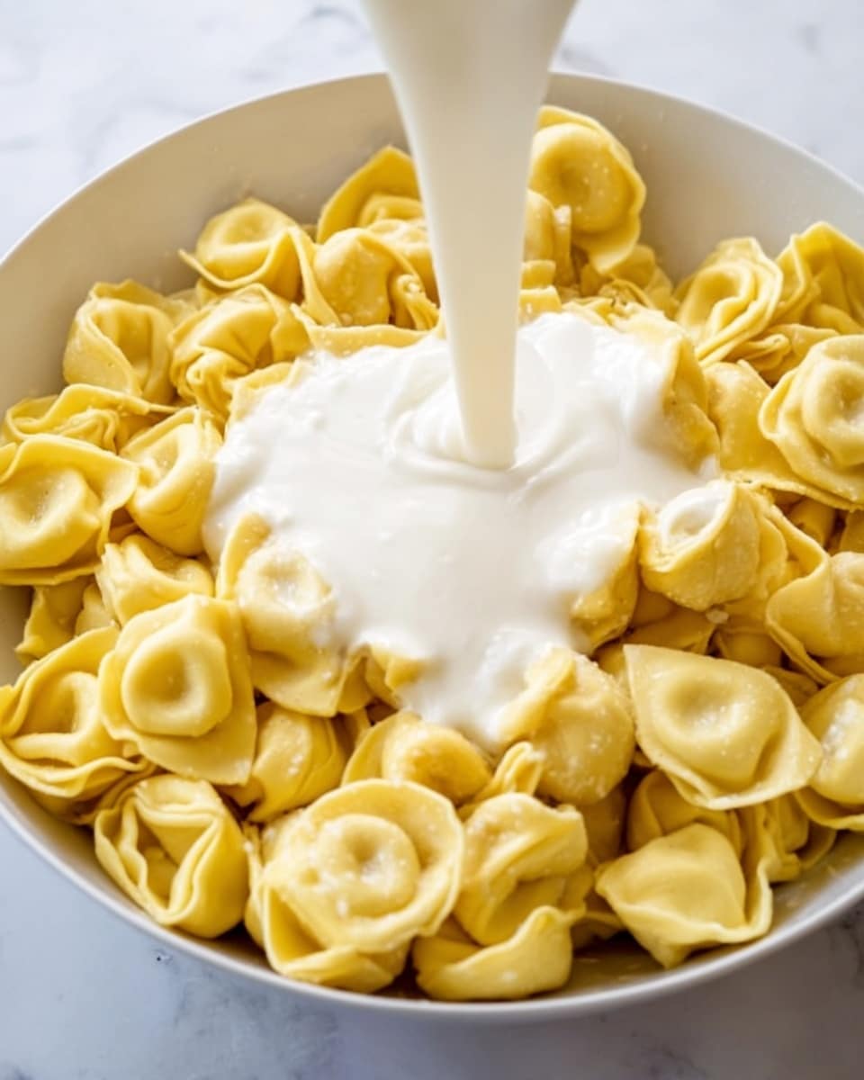 A white bowl filled with yellow tortellini pasta arranged in a circle, showing their folded round shapes with a soft texture. A creamy white sauce is being poured over the center of the pasta from above, flowing smoothly and beginning to cover some of the tortellini. The background is a white marbled surface. photo taken with an iphone --ar 4:5 --v 7