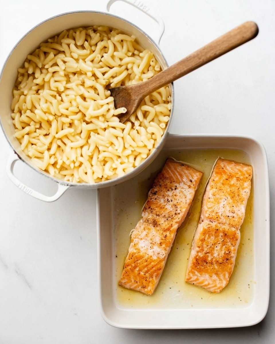 The image shows two white containers side by side on a white marbled surface. On the left, there is a white saucepan filled with cooked pasta, pale yellow in color, with soft, tubular shapes. A wooden spoon is resting inside the pan, partially submerged in the pasta, the spoon handle extending diagonally toward the top right. On the right, a white rectangular baking dish holds two cooked salmon fillets placed parallel, both light orange with browned edges and a slightly crispy texture, sitting in a shallow pool of cooking juices. Photo taken with an iphone --ar 4:5 --v 7