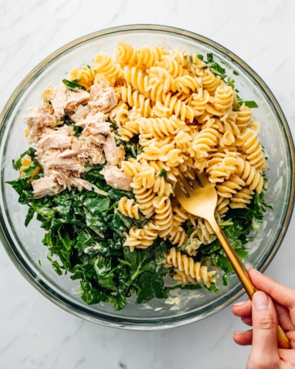 A clear glass bowl filled with a pasta salad consisting of three main layers: at the bottom, green chopped leafy vegetables, in the middle, light beige pieces of chicken or tuna, and on top, spiral-shaped fusilli pasta with a light yellow color. A woman's hand is holding a golden fork, mixing the ingredients, with the bowl placed on a white marbled surface. photo taken with an iphone --ar 4:5 --v 7