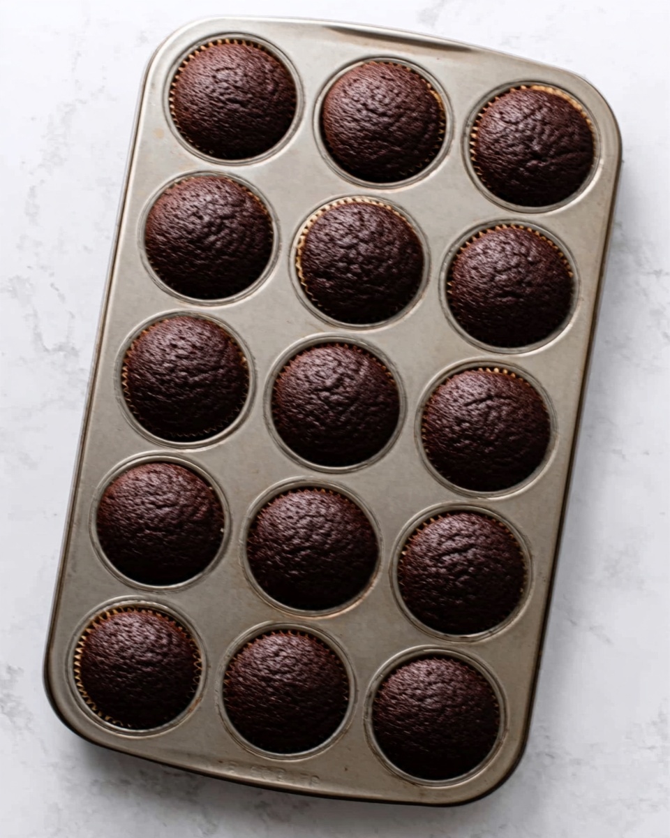 The image shows a metal muffin tray with twelve evenly spaced round slots, each filled with a fully baked dark chocolate muffin. The muffins have a rough, cracked surface texture, indicating a firm crust. The tray is placed on a white marbled surface, and in the top right corner, a woman's hand with a blue-painted fingernail is gently holding the edge of the tray. Photo taken with an iphone --ar 4:5 --v 7