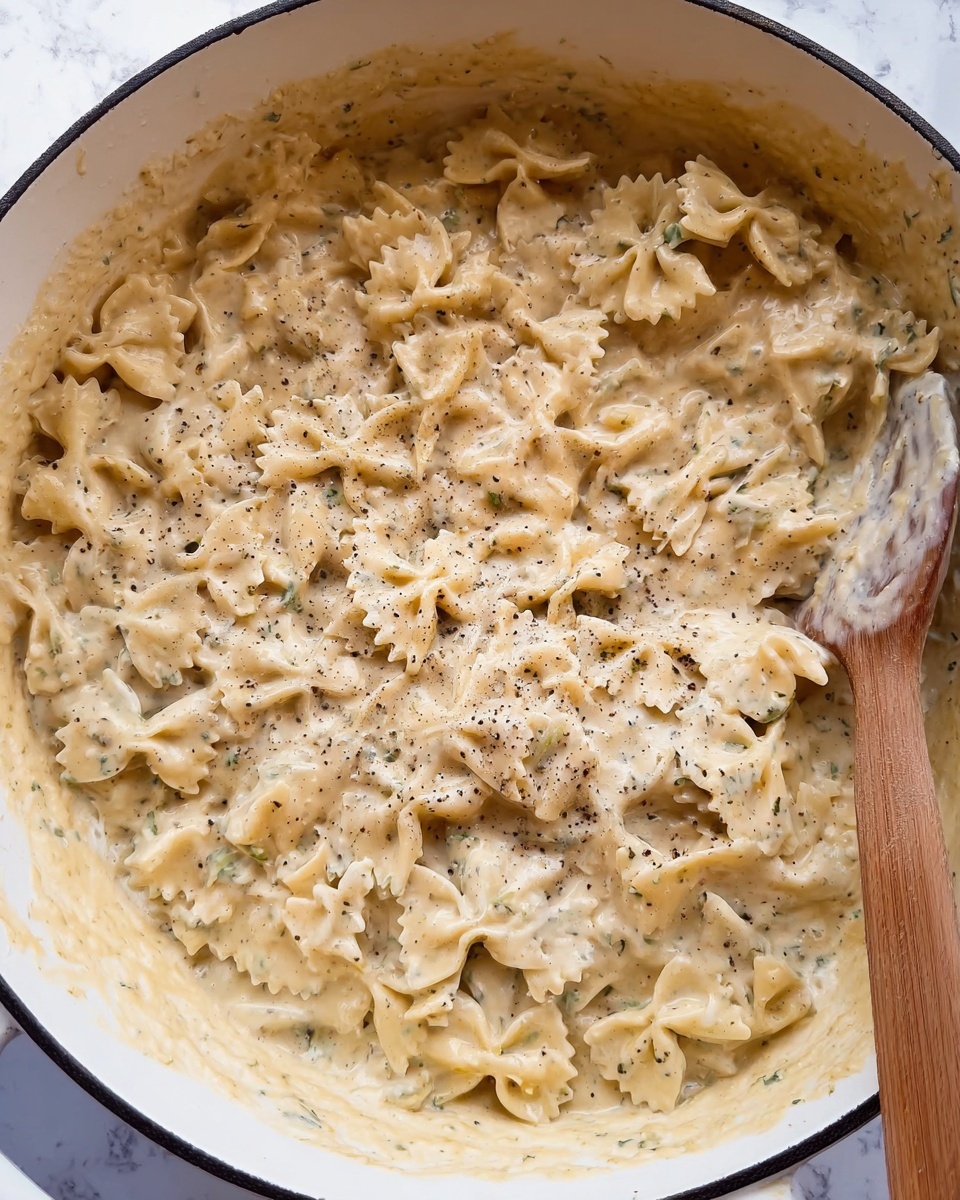 The image shows a large white pot filled with bow-tie pasta covered in thick, creamy sauce. The sauce is pale yellow with visible specks of black pepper and herbs, giving it a slightly textured look. The pasta is fully coated, with some pieces slightly sticking up, showing their ridged edges and shapes. A wooden spoon is partly dipped into the pot on the right side, mixed with the pasta but not fully submerged. The background is a white marbled surface. photo taken with an iphone --ar 4:5 --v 7
