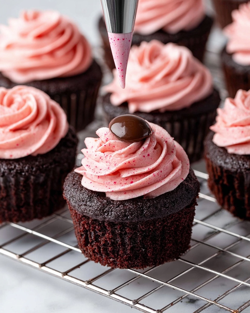 The image shows dark chocolate cupcakes on a white marbled surface with a metal cooling rack. Each cupcake has a thick, moist, rich dark brown cake base with a slightly glossy top layer. One cupcake in the front has a small dollop of shiny chocolate filling in the center. The other cupcakes have pink frosting being piped on them, with the frosting swirled in thick, soft, and fluffy layers that form a rose-like pattern on top. The pink frosting has small darker pink specks throughout, giving it a textured look. A woman's hand is holding the piping bag in the middle of adding more frosting to one cupcake. The image looks clear and bright, shot closely to show details. Photo taken with an iphone --ar 4:5 --v 7