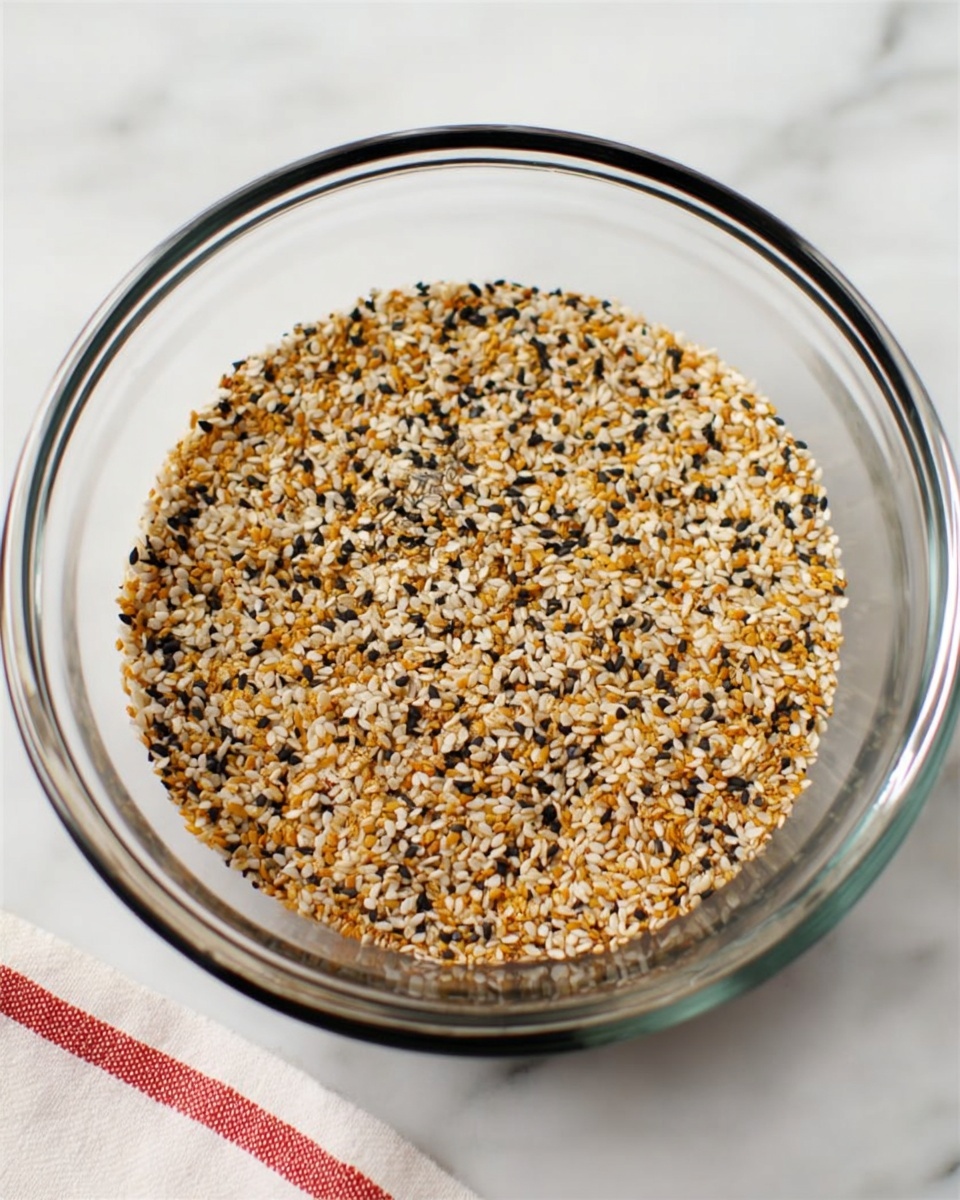 A clear glass bowl holds a single layer of a mixed seed and spice blend. The mixture shows small pieces in light golden brown, white sesame seeds, black sesame seeds, and some light yellow bits, all evenly spread out. The bowl is placed on a white marbled surface with a bit of a red and white striped cloth visible at the bottom edge of the image. The overall look is clean and simple, with the focus on the colorful mix inside the bowl. photo taken with an iphone --ar 4:5 --v 7