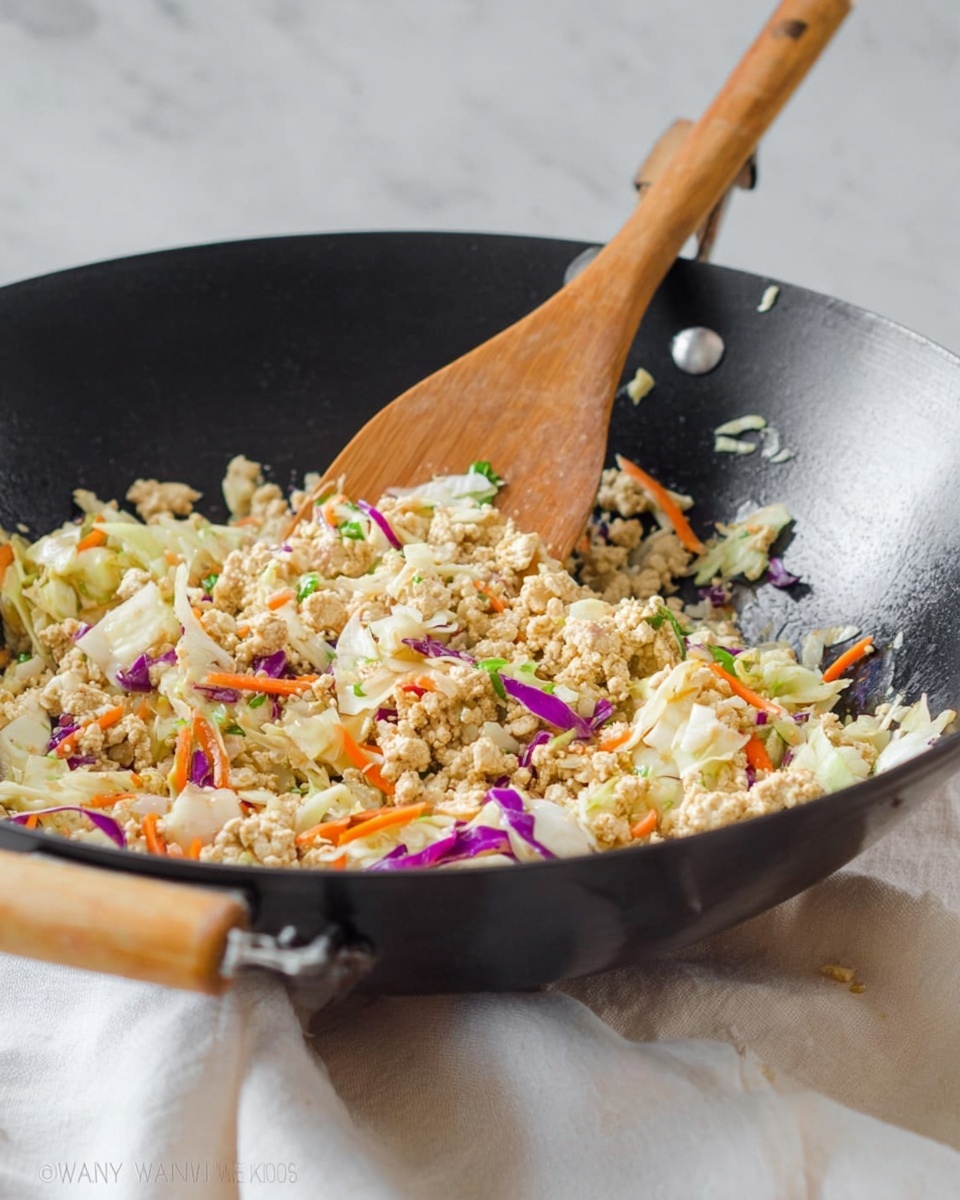 A black wok pan with light wood handles holds a stir fry mixture made of finely crumbled light tan tofu, shredded white cabbage, thin strips of bright orange carrot, and small pieces of purple cabbage, all lightly mixed together. A wooden spatula rests inside the wok, partially lifting the ingredients, showing a texture that is soft and slightly moist with small flecks of green herbs. The pan sits on a soft white cloth on a white marbled surface, with a blurred pale background. photo taken with an iphone --ar 4:5 --v 7