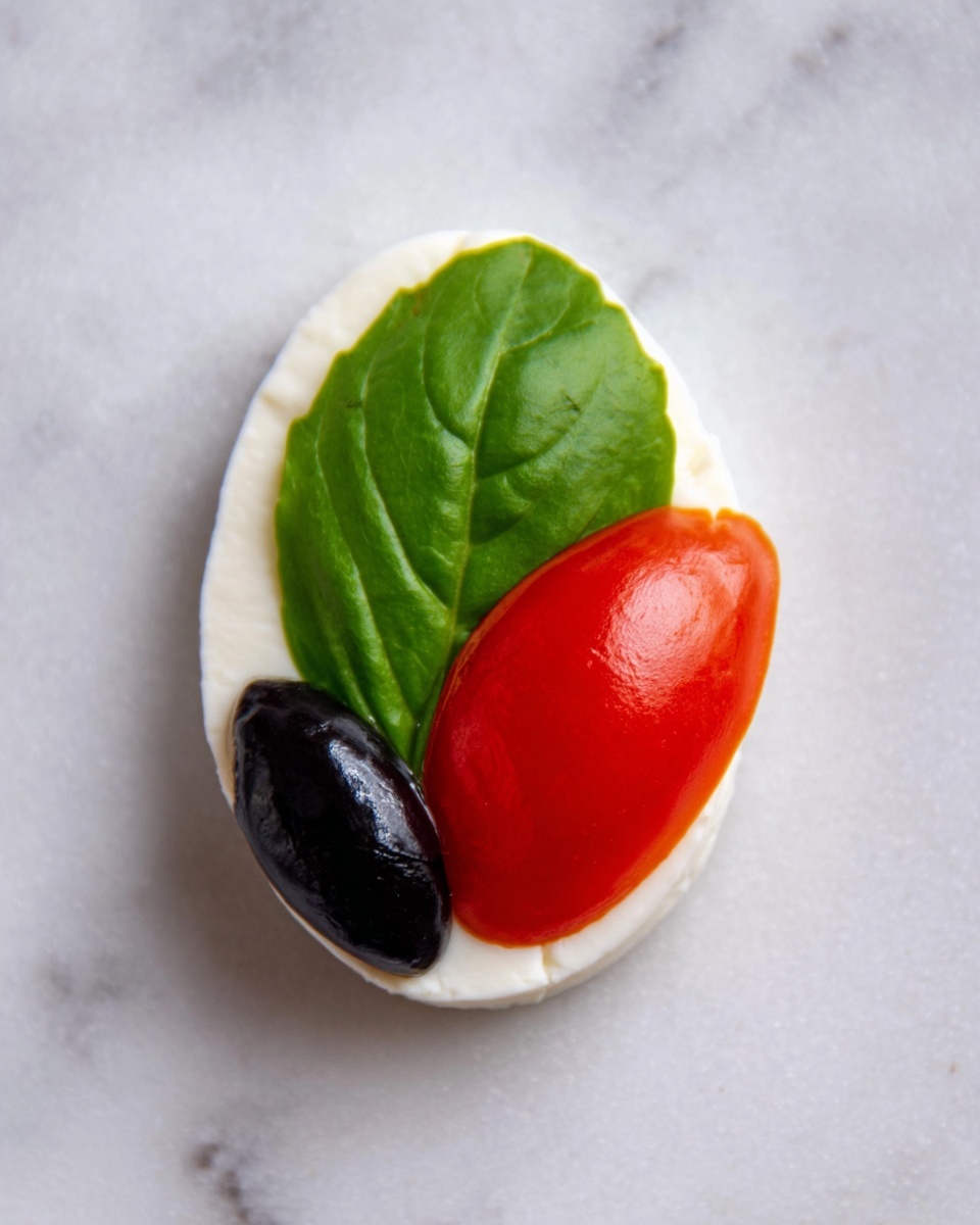 A close-up top view of a small appetizer arranged to look like a ladybug on a white marbled surface. The base layer is a white oval slice of soft cheese. On top of that, there is a fresh green basil leaf positioned lengthwise. Above the leaf, there is a whole black olive placed at one end, and a halved red grape tomato with a small wedge removed, positioned next to the olive to resemble ladybug wings. The colors are bright and clear, showing the smooth texture of the cheese, the glossy green leaf, the shiny black olive, and the vibrant red tomato. Photo taken with an iphone --ar 4:5 --v 7