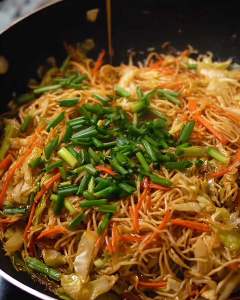 The image shows a close-up of cooked noodles mixed with shredded carrots and chopped cabbage in a black pan. On top of the noodles, there is a layer of fresh chopped green onions scattered evenly. The noodles have a light brown color with a slightly shiny texture from the sauce, while the vegetables add splashes of orange and pale green. The dish looks warm and freshly cooked with a small amount of sauce being poured over it from above. Photo taken with an iphone --ar 4:5 --v 7
