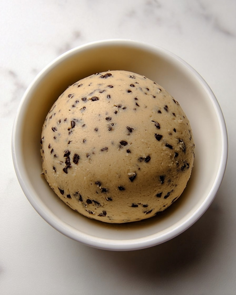 A smooth, round ball of cookie dough with small dark chocolate chips evenly spread inside is placed in a white bowl. The dough has a light beige color with the dark chocolate pieces giving a speckled look. The bowl sits on a white marbled surface, and the dough looks soft and ready to bake. photo taken with an iphone --ar 4:5 --v 7