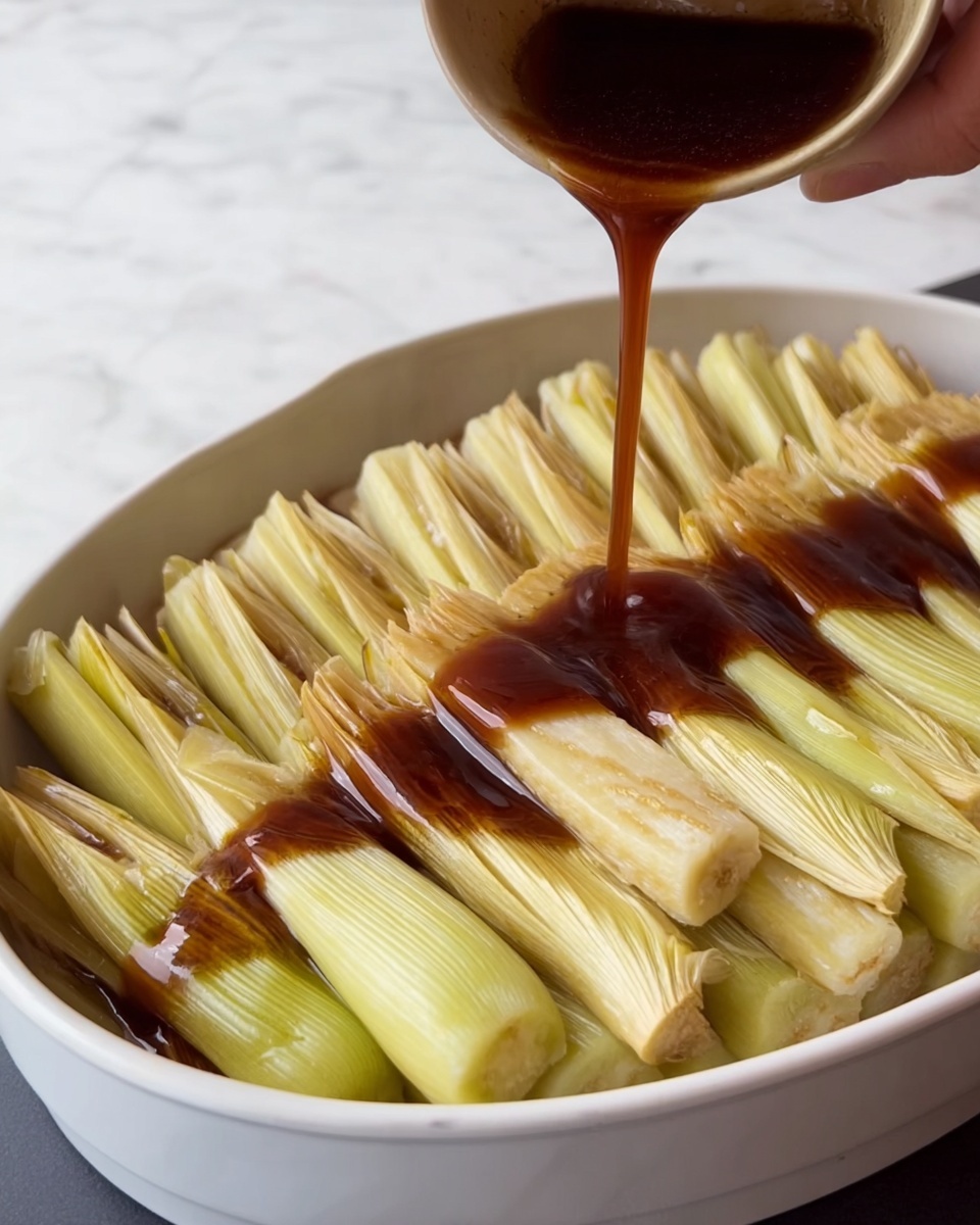 A white deep dish is filled with multiple folded greenish-yellow layers of bamboo shoots standing upright in a neat arrangement, each piece showing a smooth and slightly ribbed texture with a pale inner color. A dark brown sauce is being poured over the layers, pooling at the bottom and filling the gaps between the shoots, creating a glossy and rich surface. The scene is set on a white marbled surface and a woman's hand is steadying the dish while pouring the sauce. photo taken with an iphone --ar 4:5 --v 7