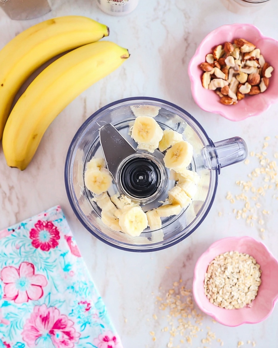The image shows a top view of a clear food processor filled with several small pieces of peeled banana placed on the sharp metal blade inside. The food processor sits on a white marbled surface. To the left side, there are two whole yellow bananas lying on a floral-patterned cloth with pink and blue flowers. On the right side, there are two small pink bowls; the top one contains brown nut pieces, and the bottom one holds a pile of beige oats. Some loose nut pieces are scattered on the white marbled surface near the pink bowls. The scene is bright and colorful, with a clean and fresh look. photo taken with an iphone --ar 4:5 --v 7