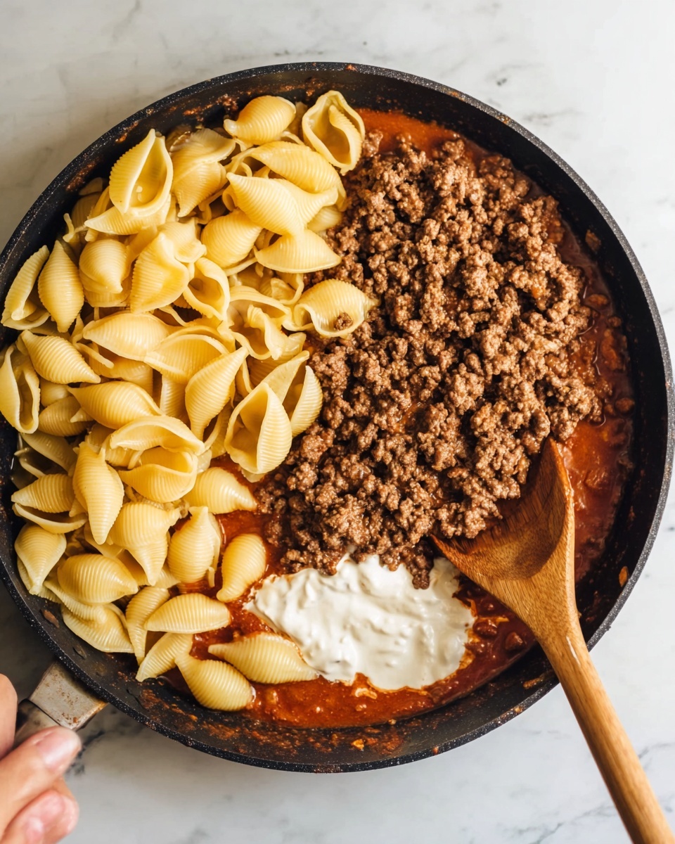 The image shows a top view of a black pan on a white marbled surface, filled with three main layers of food. On the left side, yellowish cooked shell pasta is gathered. On the right, there is a thick layer of browned ground meat over a dark red sauce, with a dollop of white creamy sauce partially mixed in near the bottom right. A large wooden spoon is resting horizontally across the pan, with a woman's hand holding the handle from the top left corner. photo taken with an iphone --ar 4:5 --v 7