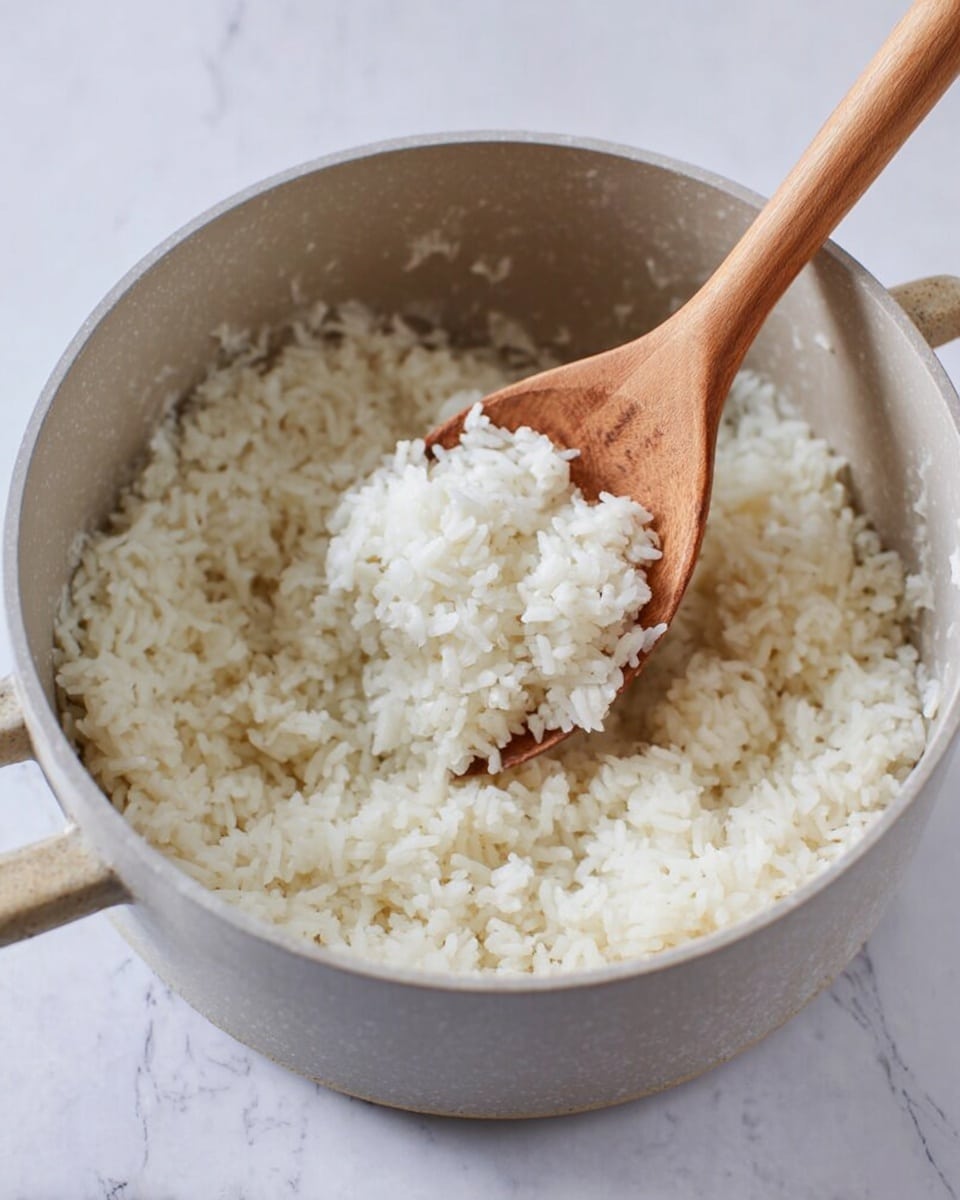 A light gray pot filled with soft, white cooked rice. A wooden spoon with a smooth texture is scooping some rice from the pot, showing small, fluffy rice grains. The pot sits on a white marbled surface with subtle gray veins. photo taken with an iphone --ar 4:5 --v 7